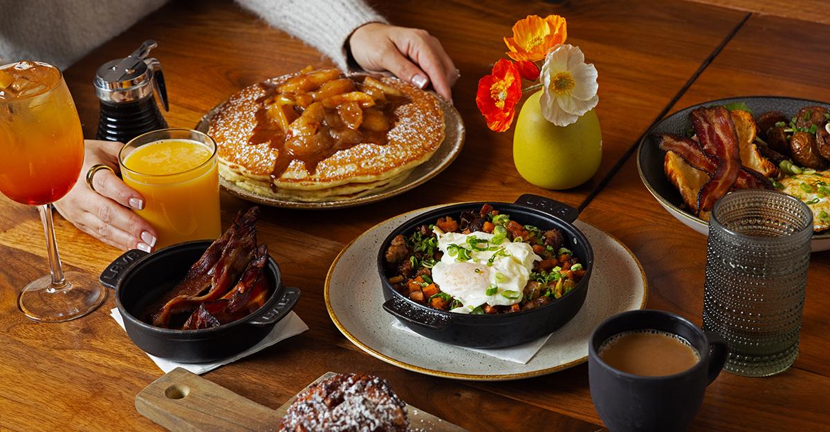 Brunch spread with pancakes, eggs, bacon, drinks, and flowers on a wooden table.
